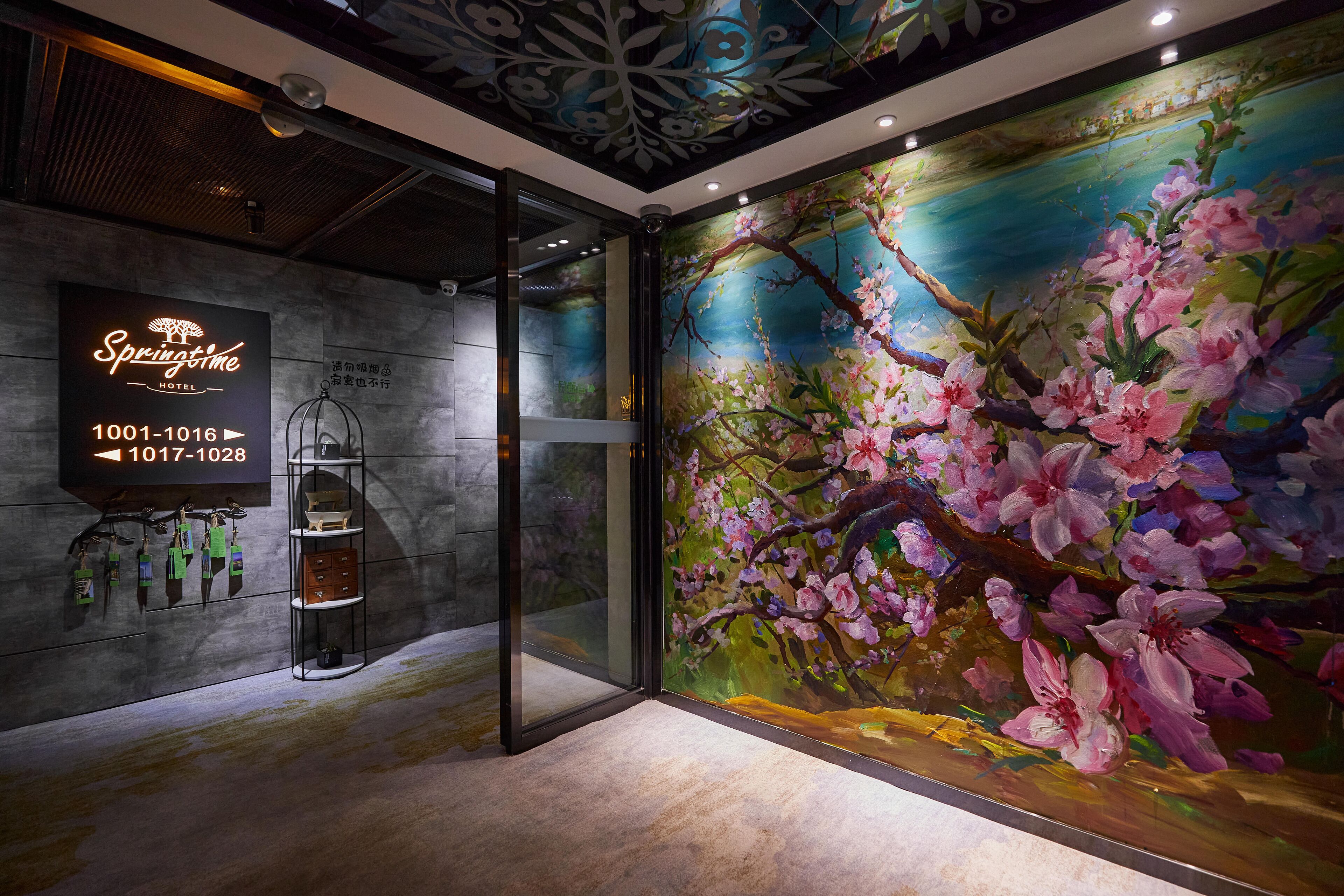 A hotel lobby with a large floral mural, a glass door entrance, and a signboard displaying room numbers.
