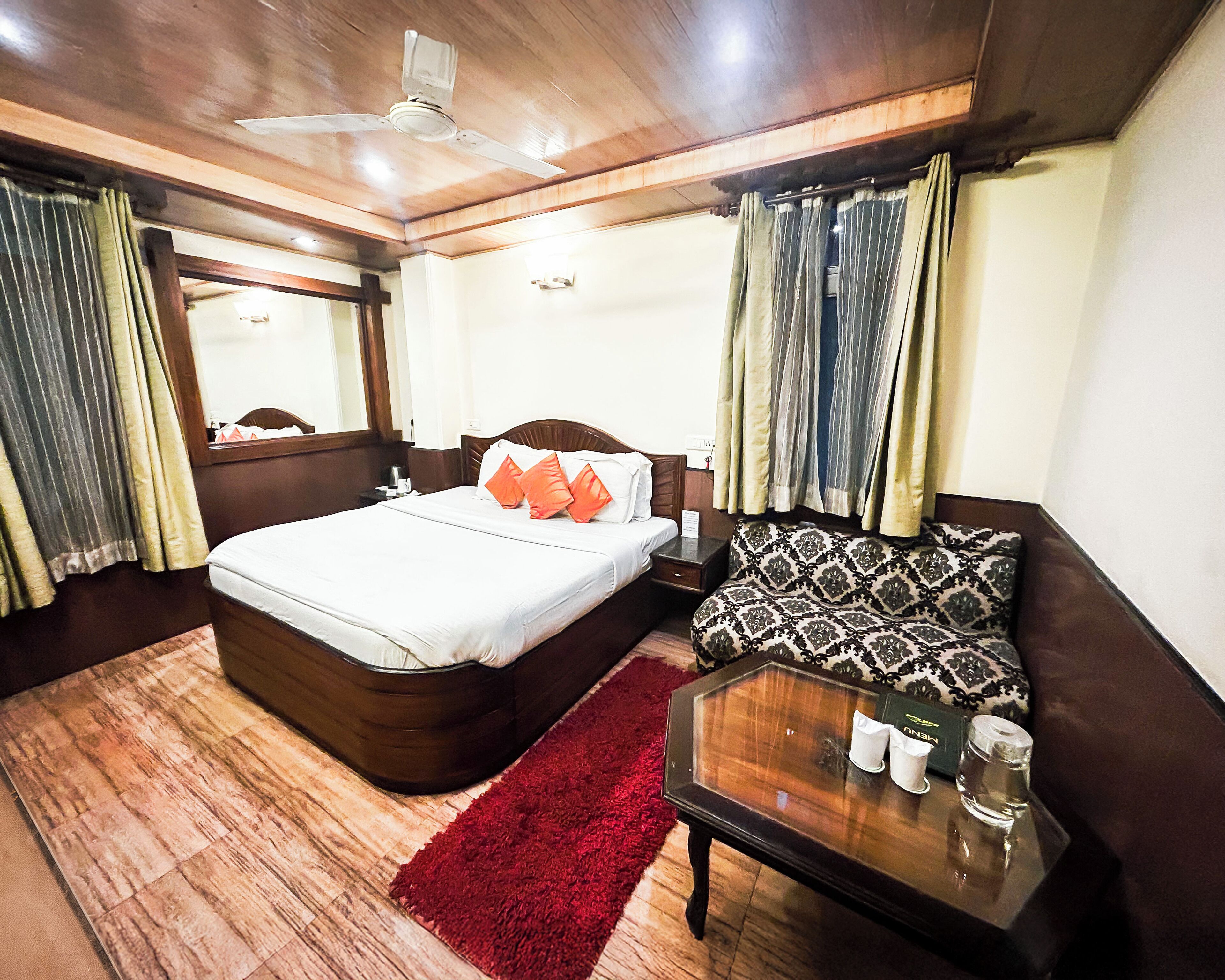 Une chambre d’hôtel avec un plafond en bois, un lit recouvert de draps blancs et agrémenté d’oreillers orange, un canapé à motifs, une table basse en verre et un tapis rouge.