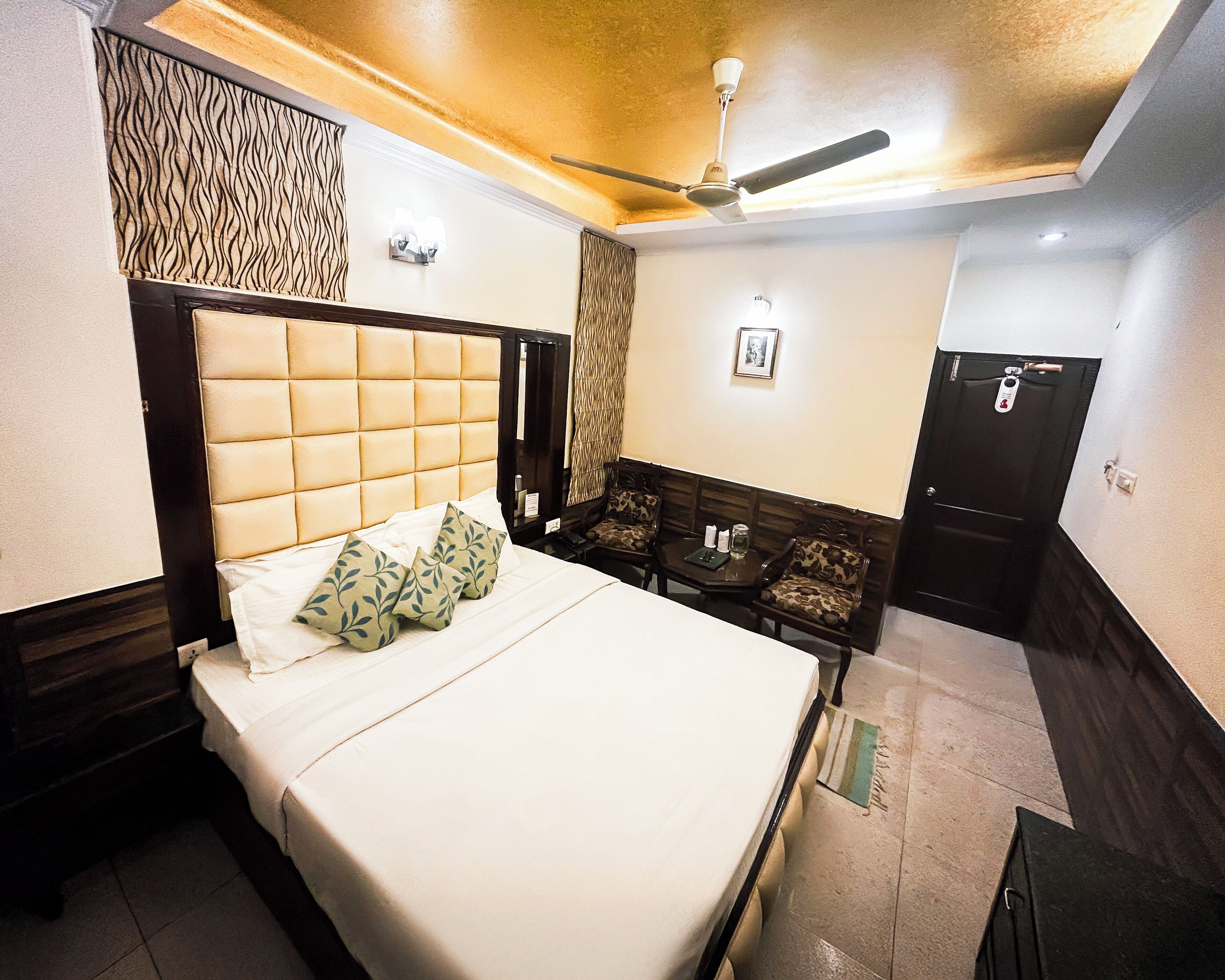 A hotel room with a bed, a ceiling fan, a small table with chairs, and a door.
