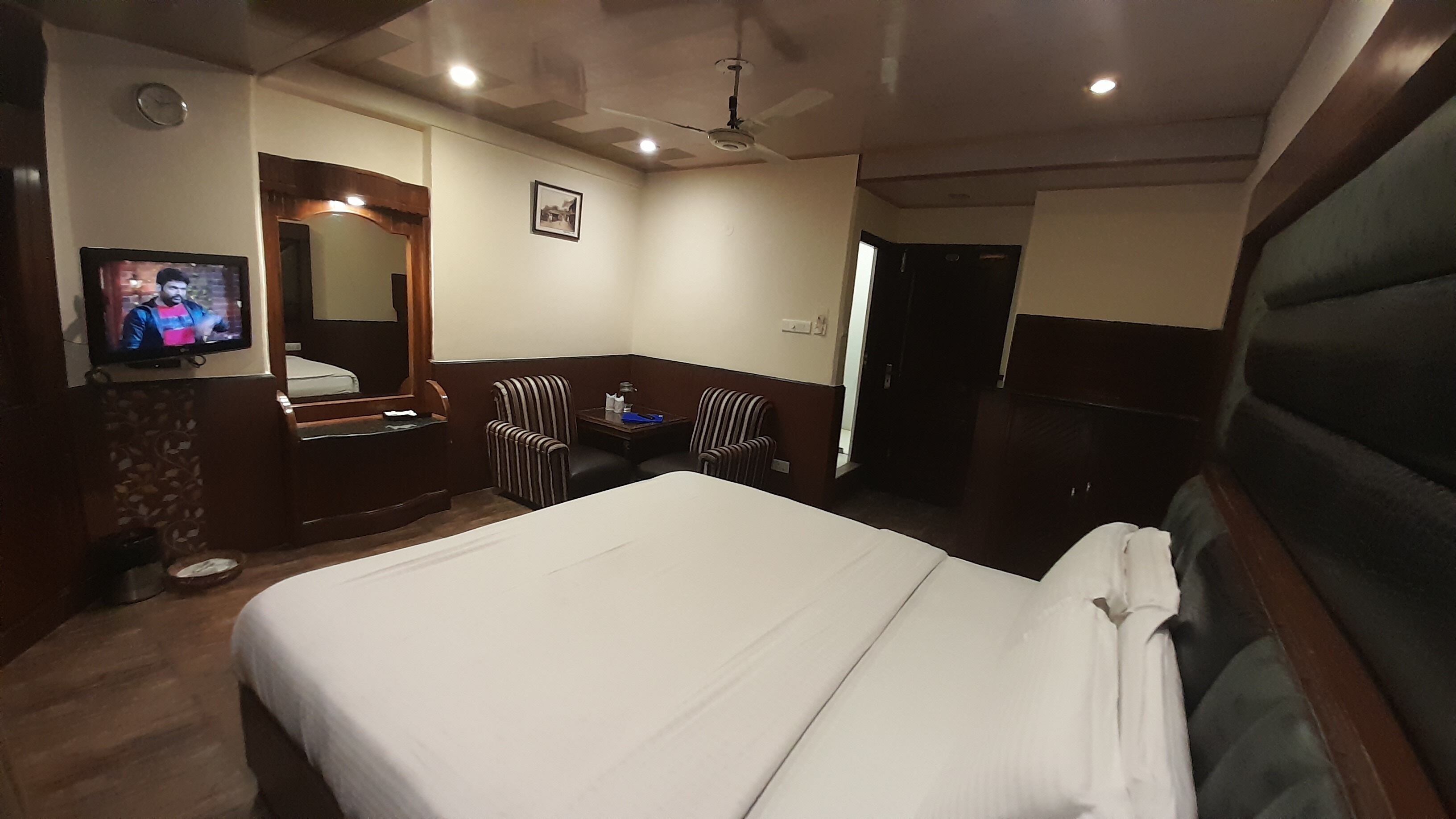 Une chambre d’hôtel équipée d’un lit, d’une télévision, d’un bureau, d’une chaise, d’un miroir et d’un ventilateur de plafond.
