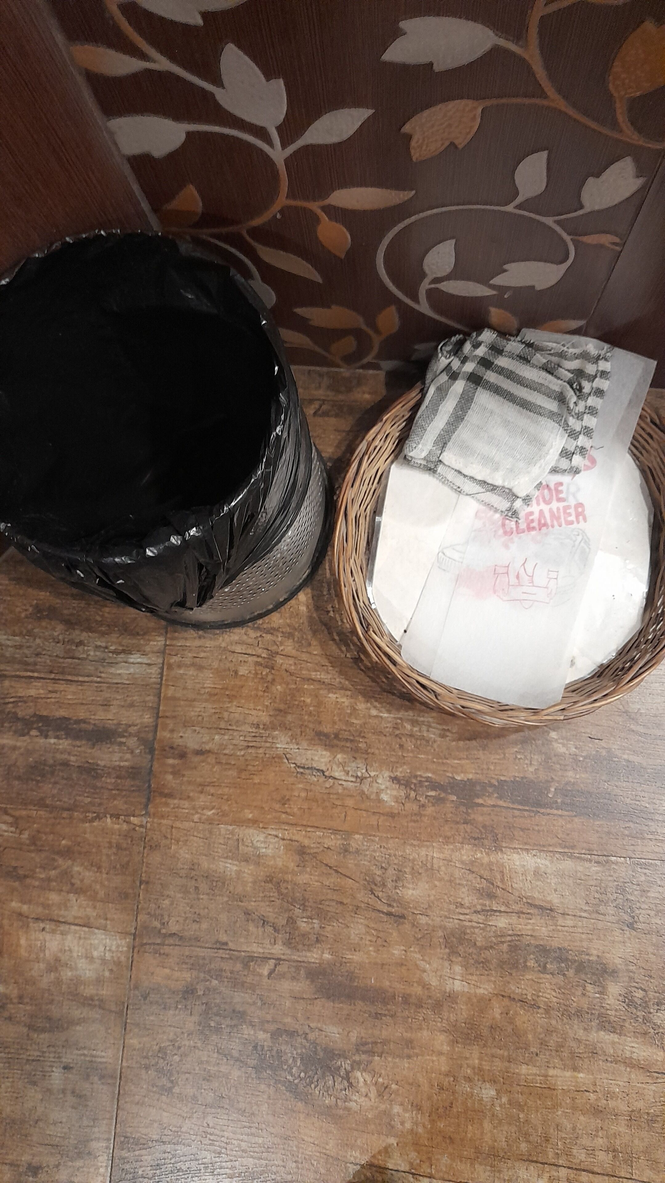 A black trash bin and a wicker basket with a white bag labeled "STORAGE CLEANER" on a wooden floor.