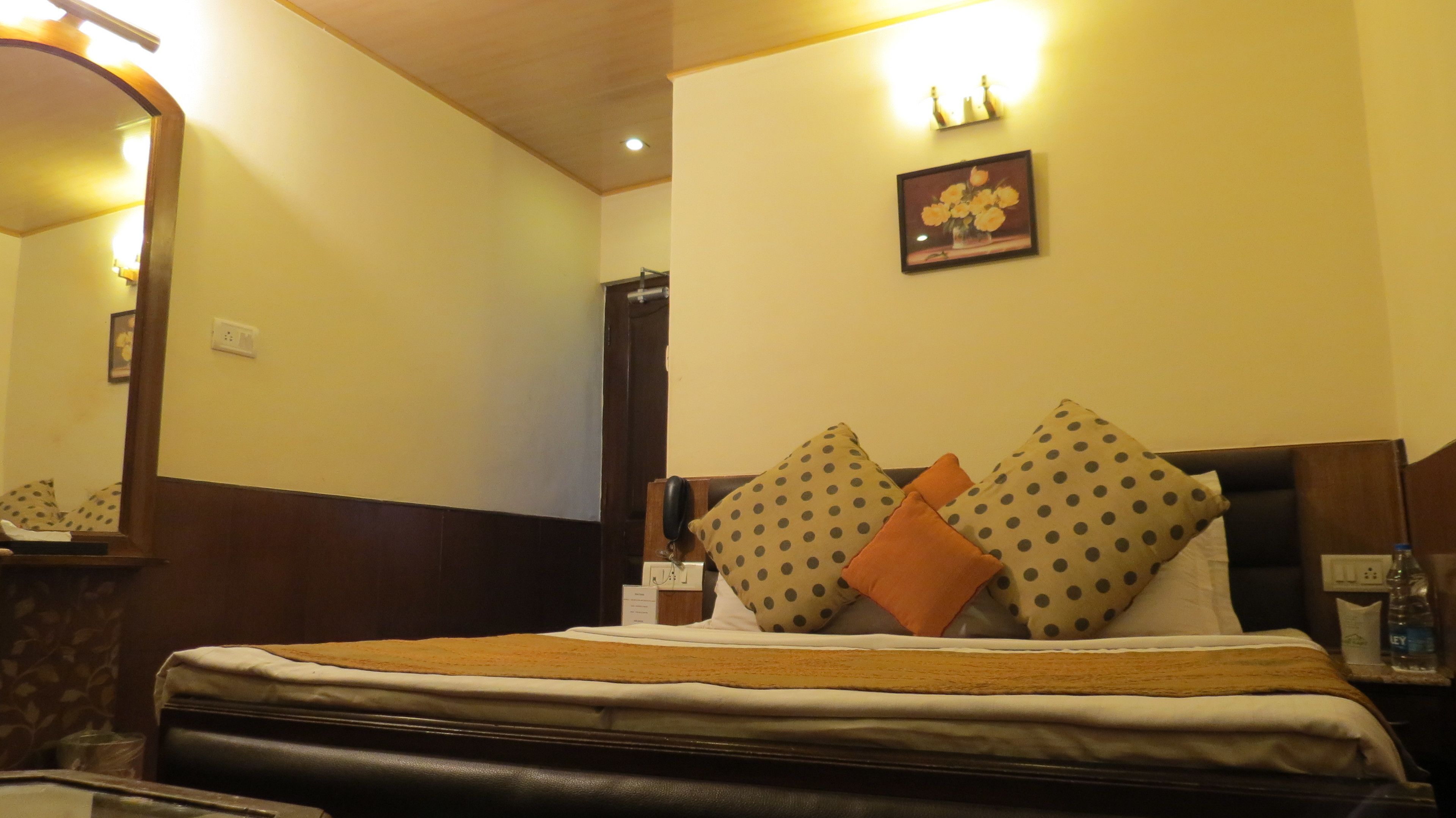 A hotel room with a bed, a bedside table, a mirror, a wall-mounted light, and a framed picture.