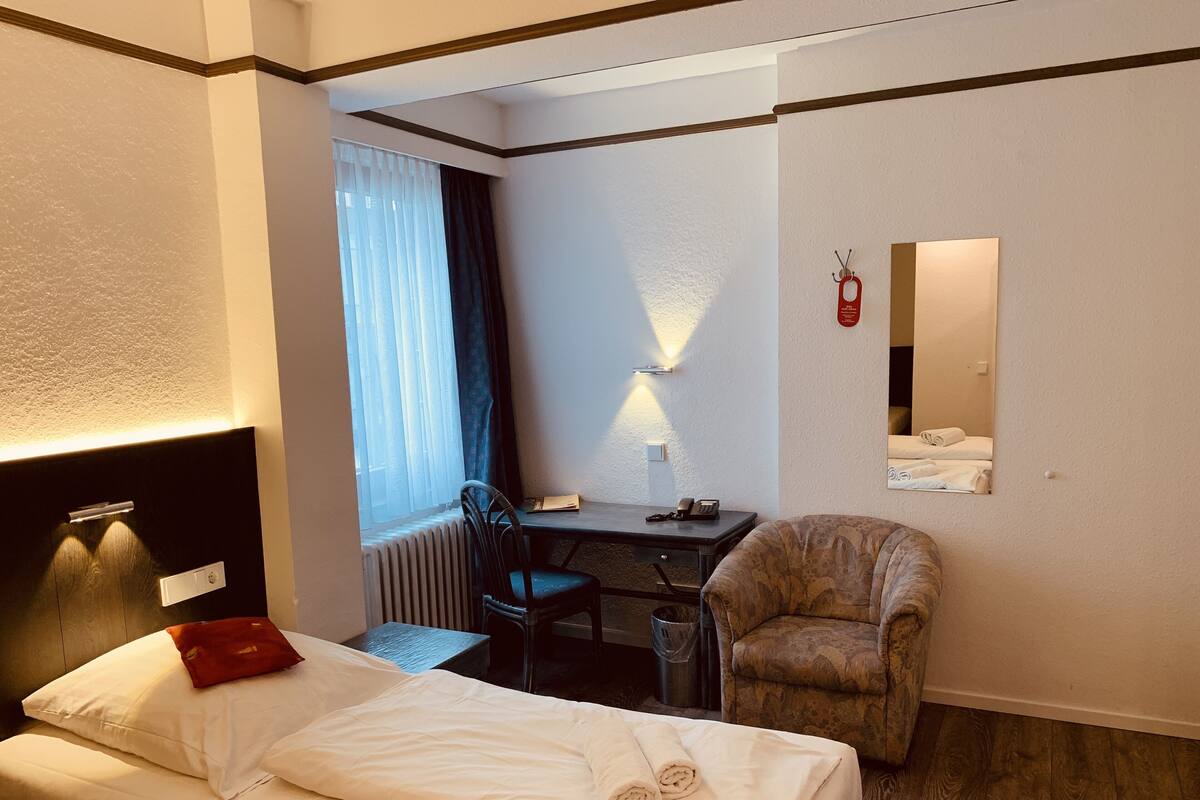 A hotel room with a bed, a desk, a chair, a radiator, and a window with curtains.