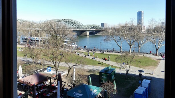 A view of a riverside park with a bridge, trees, and people walking. - Kunibert der Fiese (Cologne)