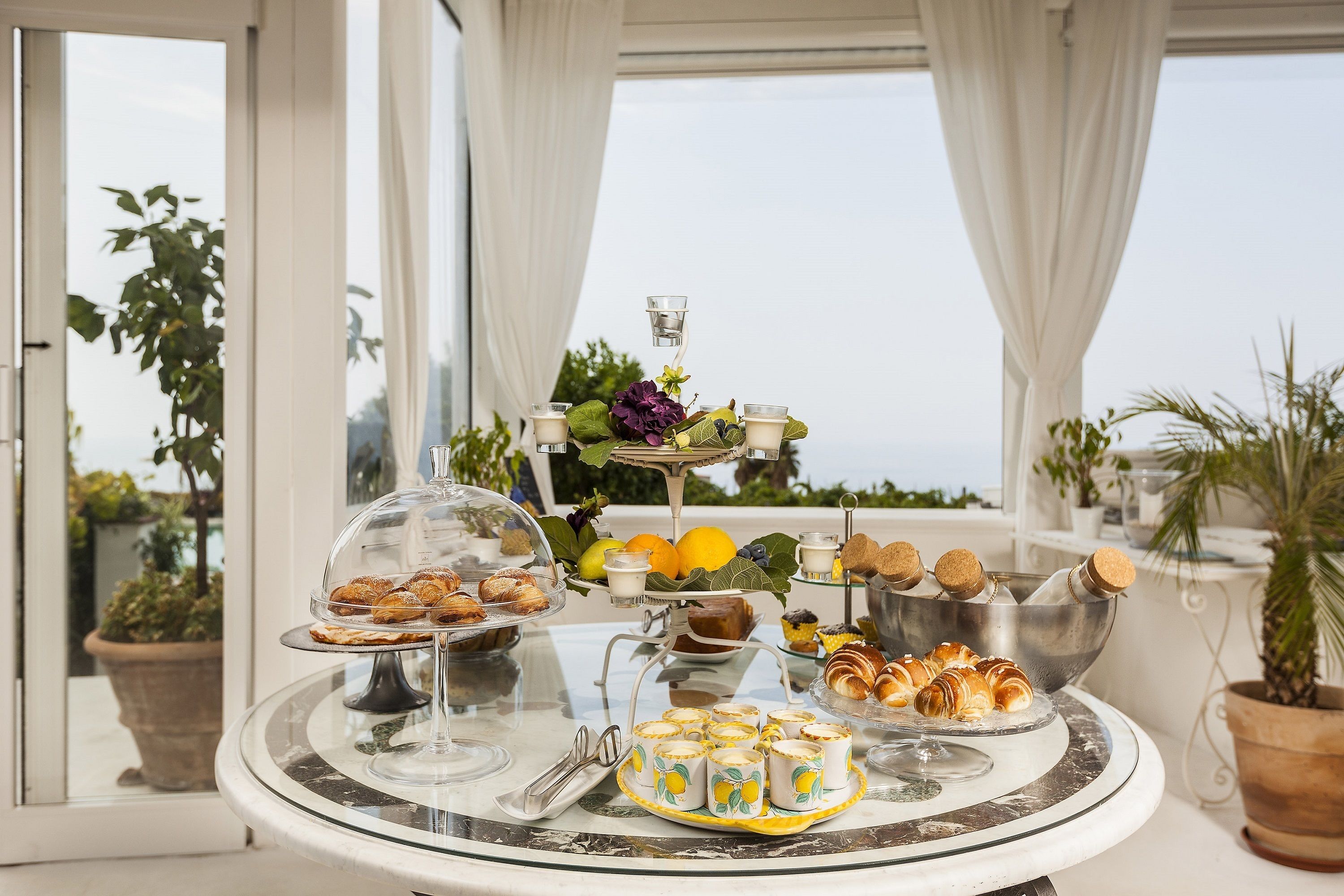A table with various pastries and drinks, set against a backdrop of large windows and potted plants.