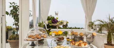 A table with various pastries and drinks, set against a backdrop of large windows and potted plants.