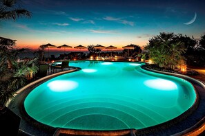 Una piscina con una luna creciente en el cielo, rodeada de palmeras y sillones de descanso.