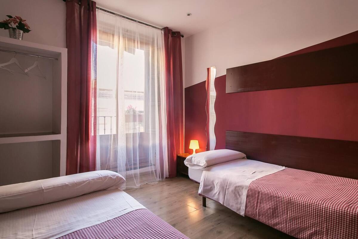 A hotel room with two beds, a large window with sheer curtains, a bedside table with a lamp, and a view of the outside.
