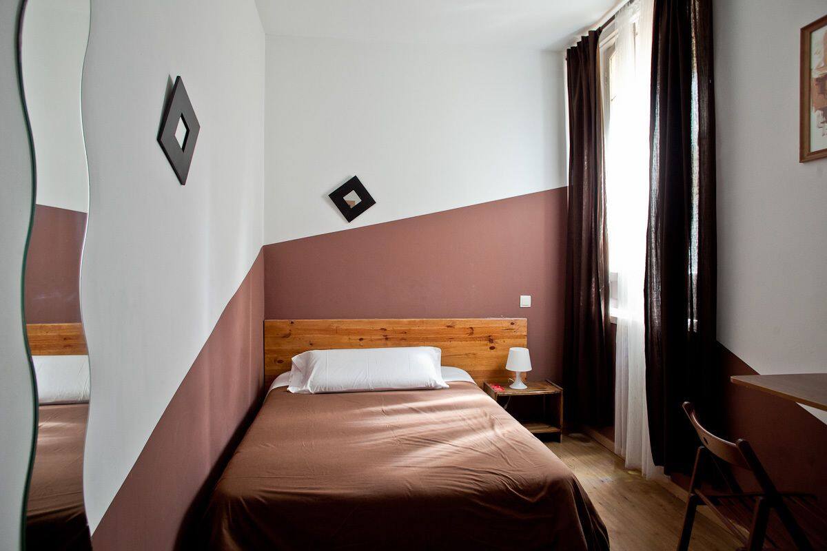 A hotel room with a wooden headboard, a single bed with a brown bedspread, a nightstand with a lamp, a chair, and a window with curtains.