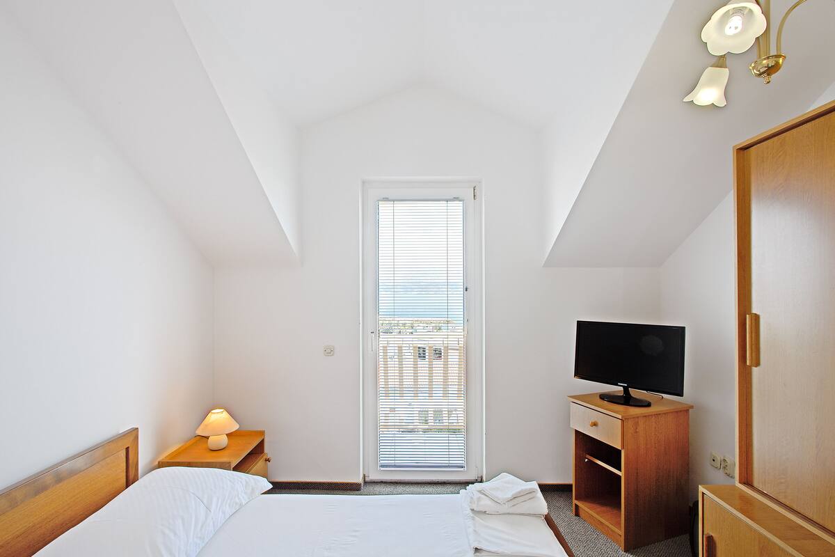 A bedroom with a bed, a TV on a wooden stand, a nightstand with a lamp, and a door leading to a balcony.