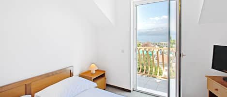 Una camera da letto con un letto, un comodino con una lampada, una TV e una porta-finestra su un balcone affacciato su edifici e alberi.