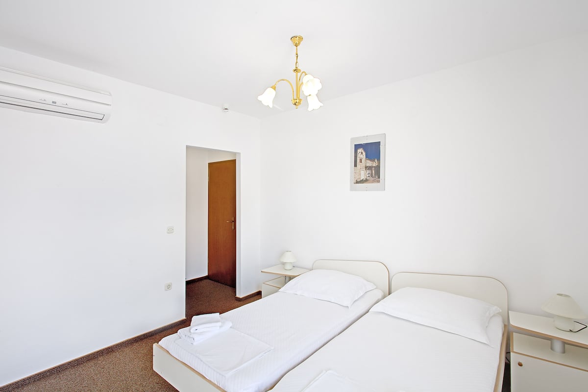 A bedroom with a bed, bedside tables, a lamp, an air conditioner, and a door leading to another room.