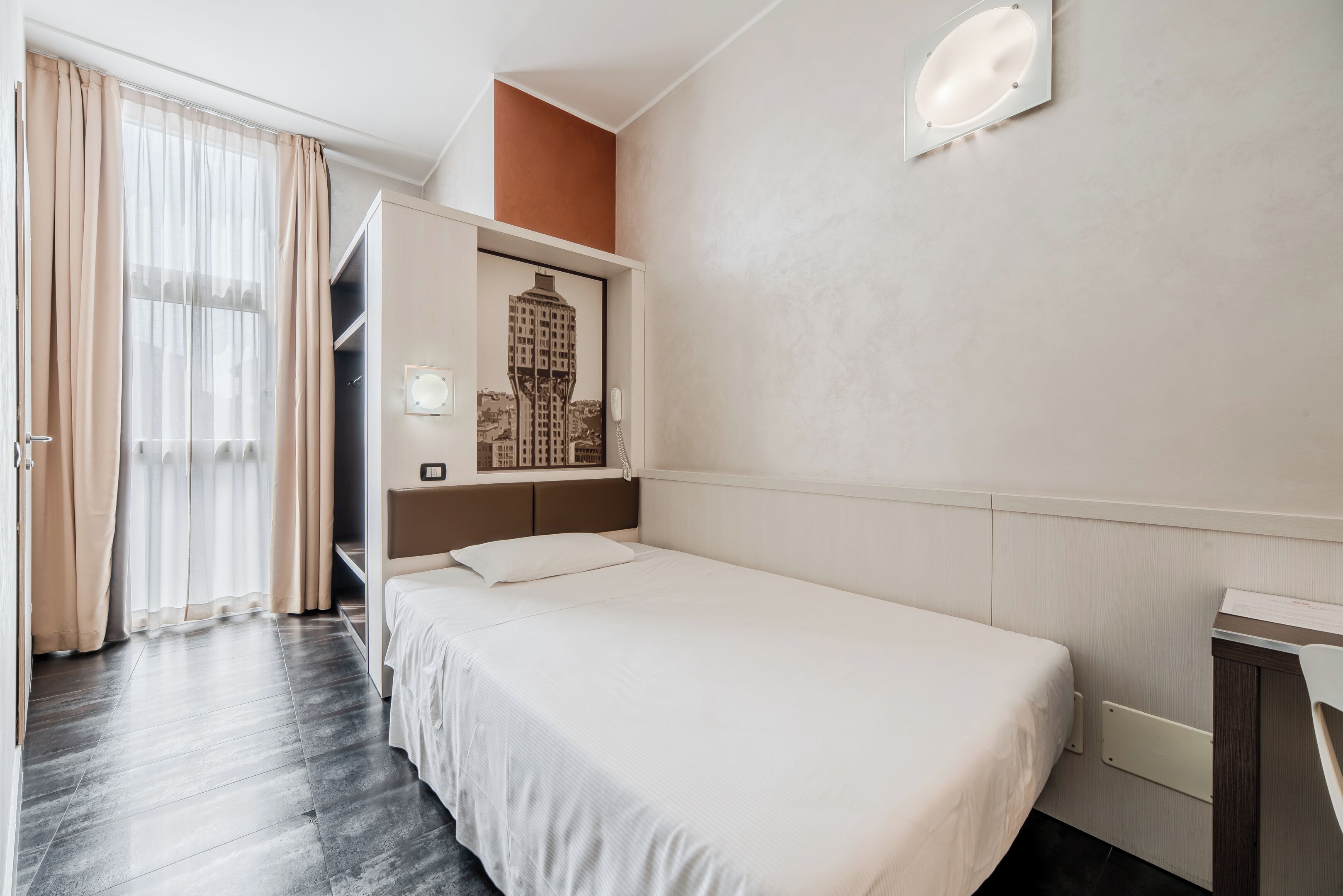 A hotel room with a single bed, a wardrobe, a small desk, and a window with curtains.