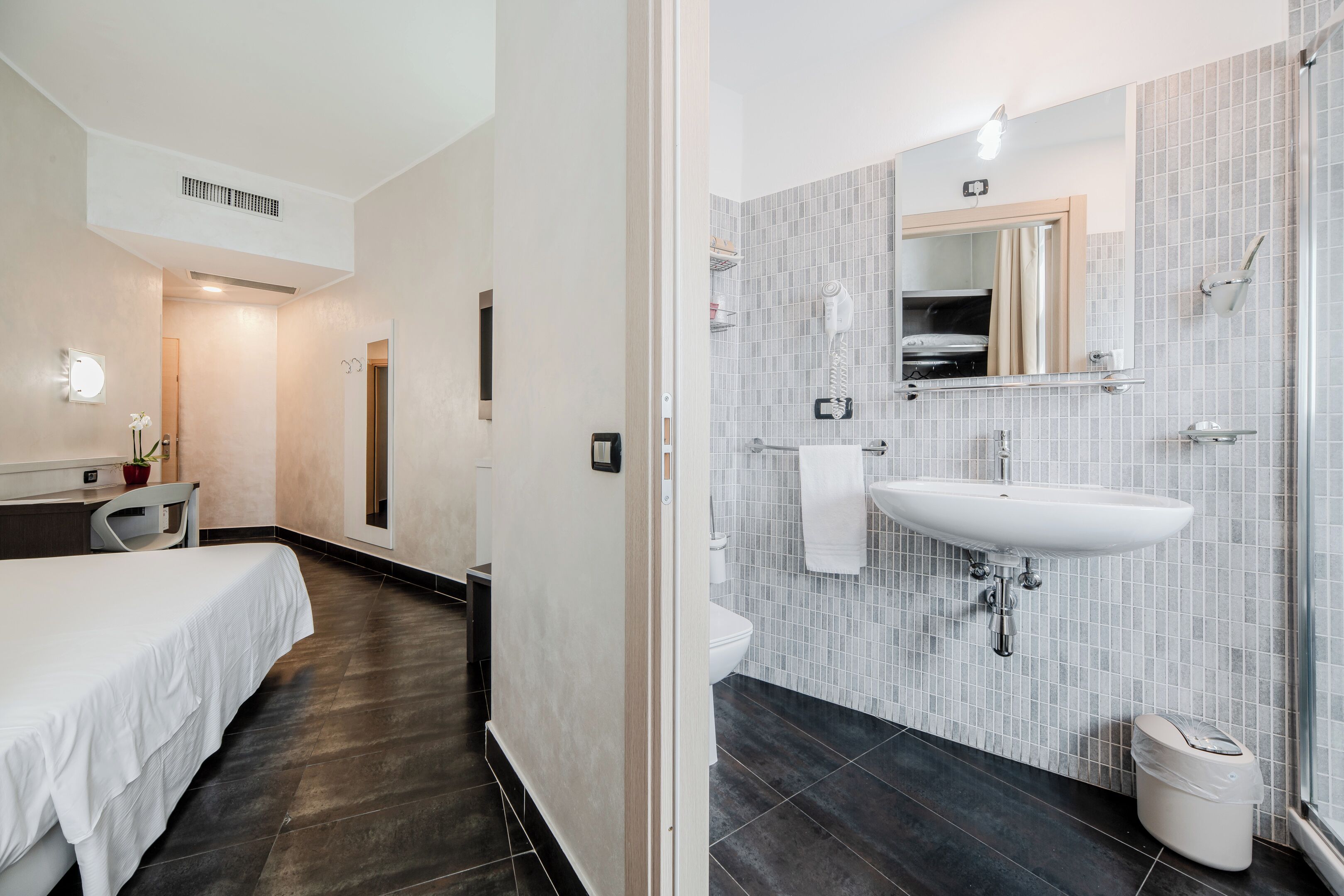 Un baño de hotel moderno con un espejo grande, un lavabo blanco y un piso de baldosas oscuras.
