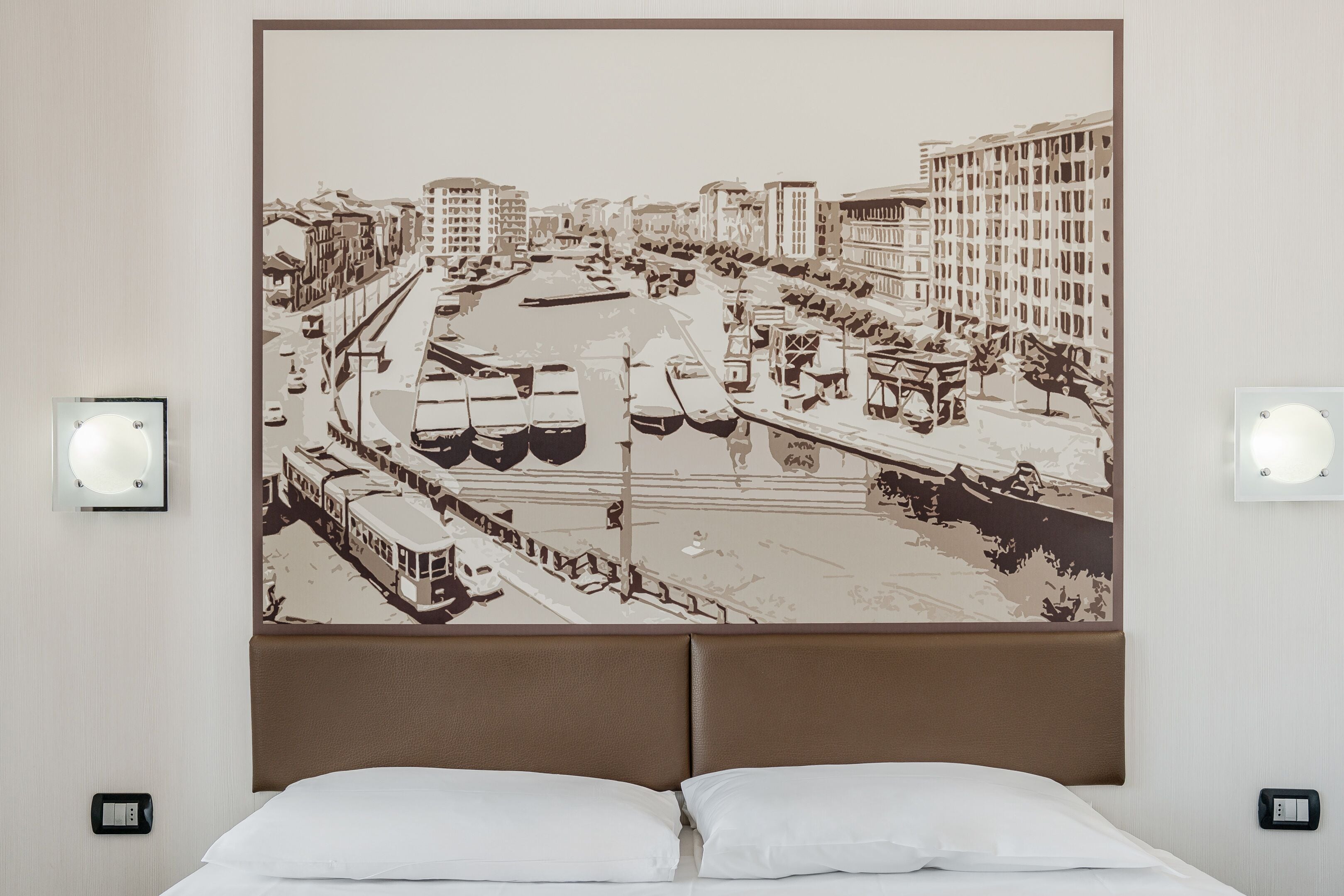 Camera d'albergo con un grande murale raffigurante un panorama cittadino, un letto con lenzuola bianche e due interruttori a parete.