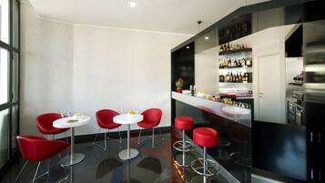 Un espace bar moderne avec des chaises et des tables rouges, un comptoir de bar où sont exposés des bouteilles de vin et de spiritueux, et un coin cuisine à l’arrière-plan.
