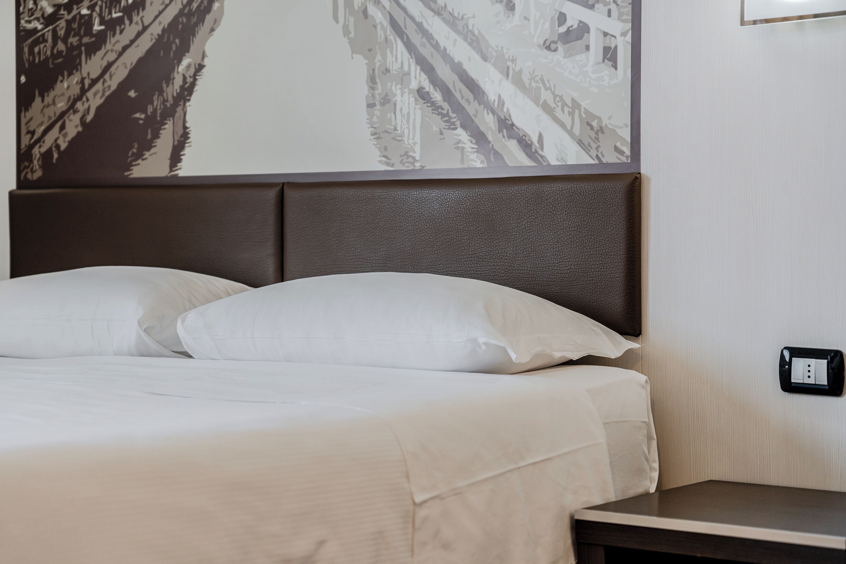 Una cama bien hecha con sábanas blancas y un cabecero oscuro, una mesita de noche y un enchufe en la pared.
