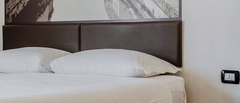 Una cama bien hecha con sábanas blancas y un cabecero oscuro, una mesita de noche y un enchufe en la pared.