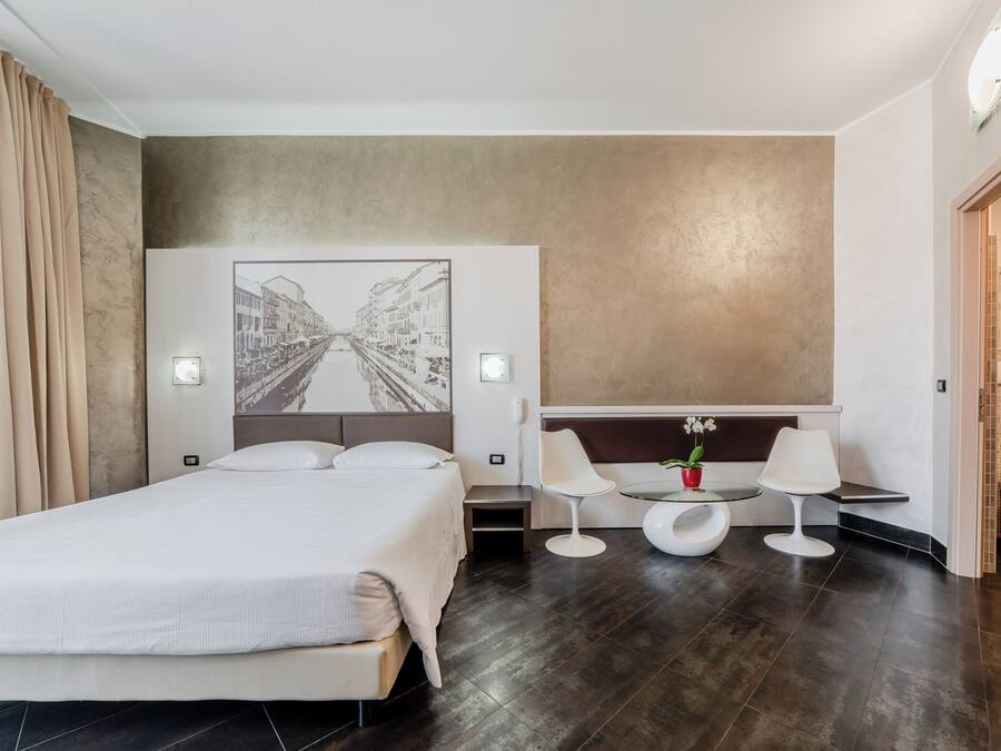 A modern hotel room with a large bed, a small round table with two chairs, a wall-mounted phone, and a large framed picture.