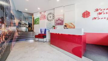 Un hall d'hôtel avec un comptoir d'accueil rouge, une horloge et un escalier menant à un étage supérieur.
