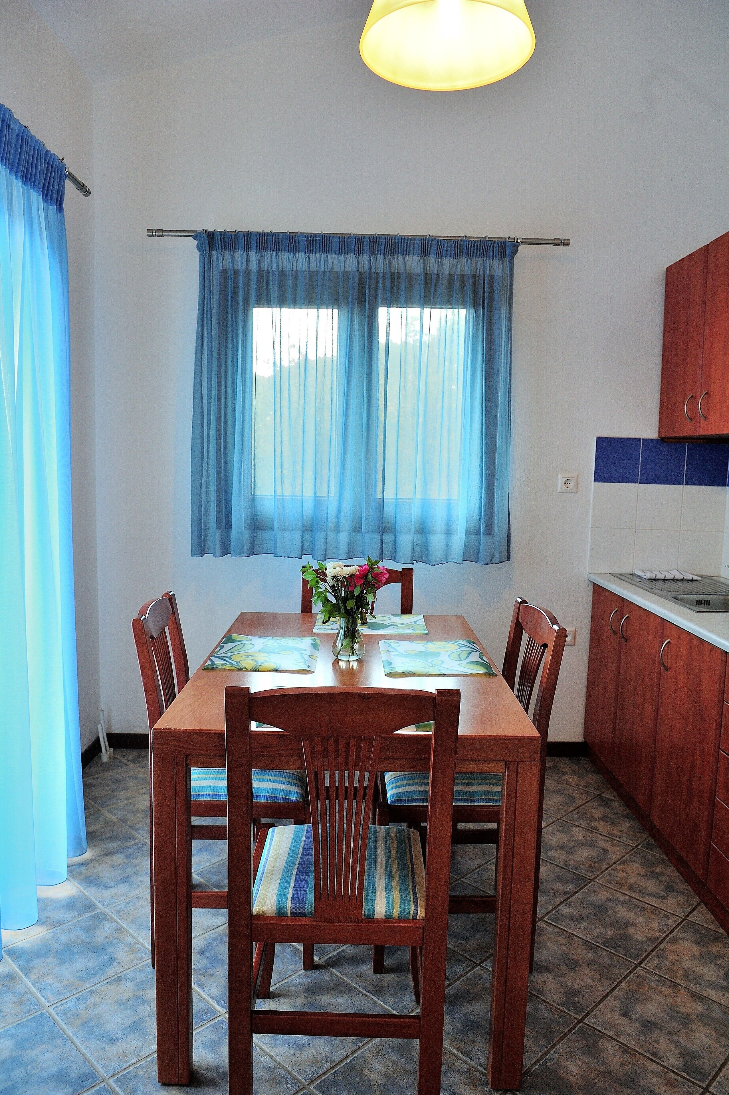 Un comedor con una mesa de madera, sillas, un florero con flores y una ventana con cortinas azules.