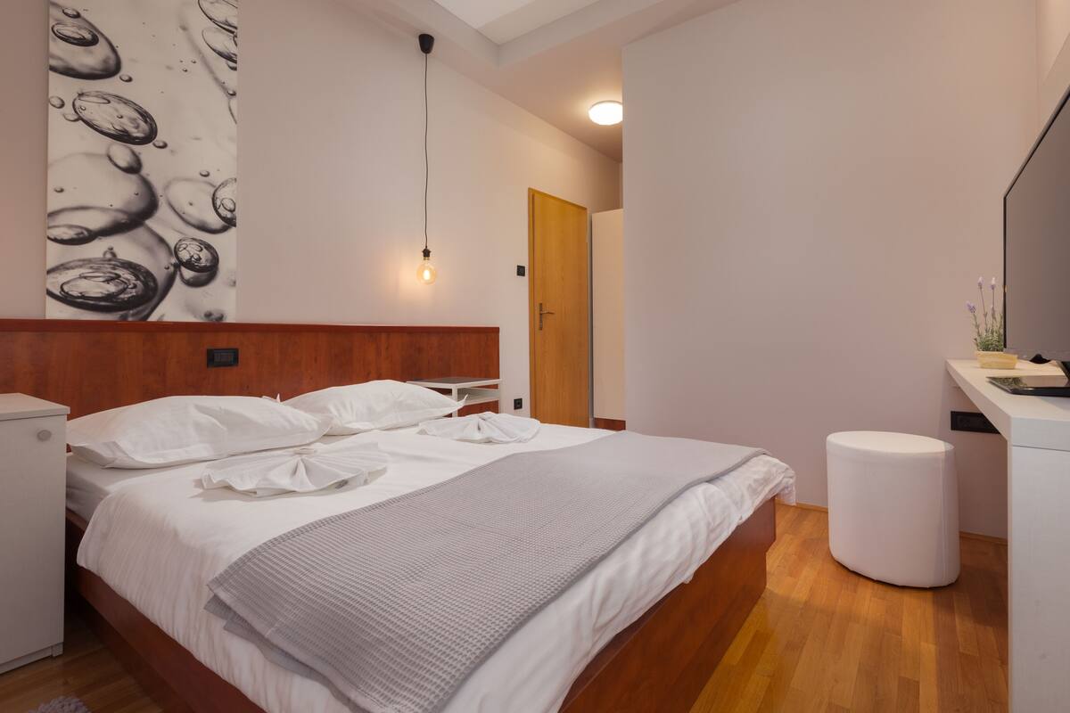 Una habitación de hotel moderna con cabecera de madera, una cama con ropa de cama blanca, una mesita de noche pequeña y blanca, un otoman blanco redondo y un televisor montado en la pared.