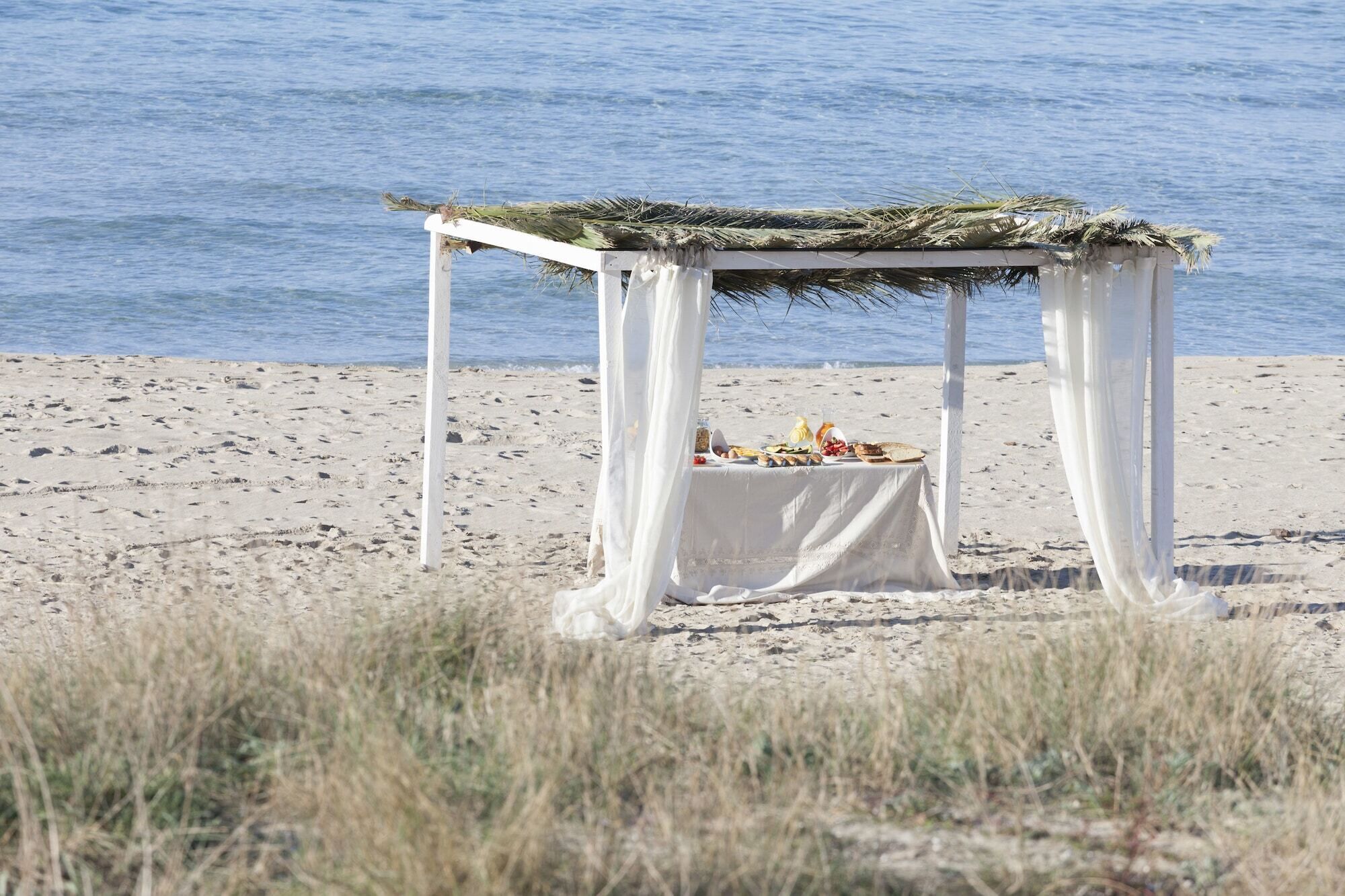 Un montaje para comer frente al mar, con una mesa cubierta con tela blanca, rodeada de pilares blancos y un techo de paja.