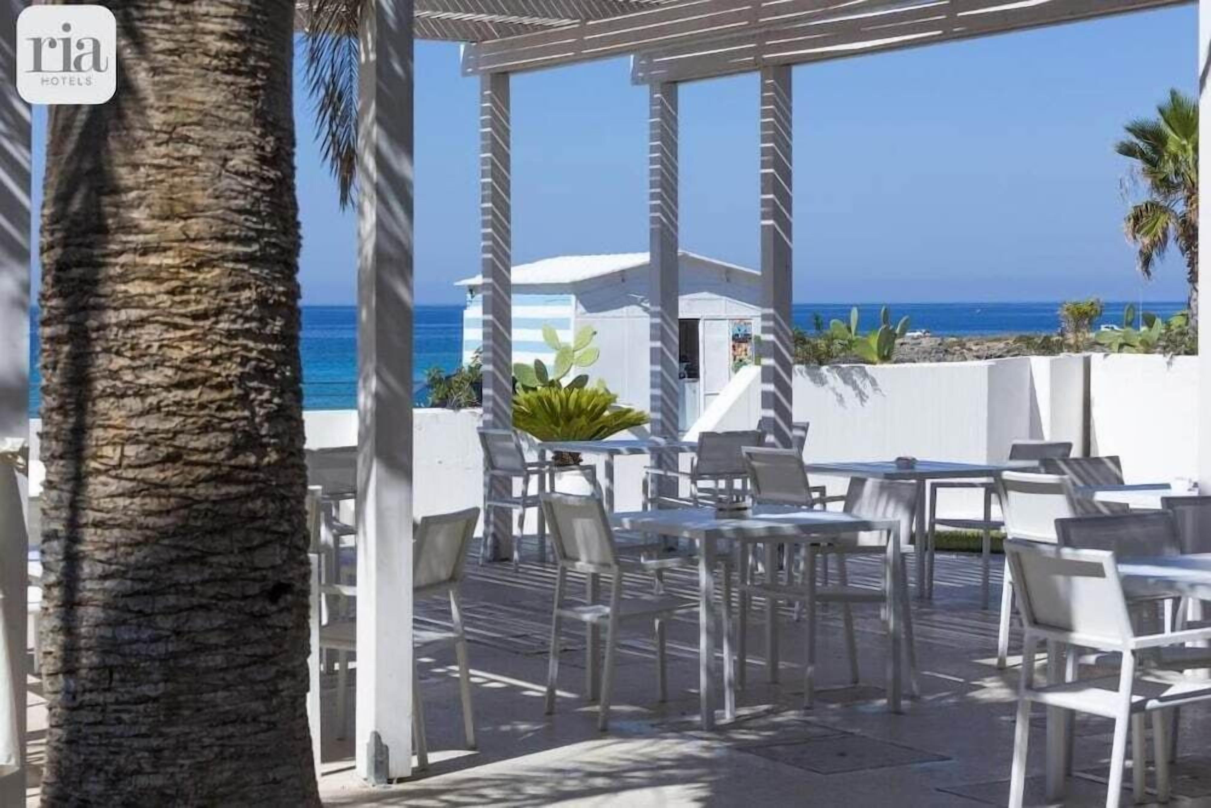 Eine Dachterrasse mit weißen Tischen und Stühlen mit Blick auf einen Strand.