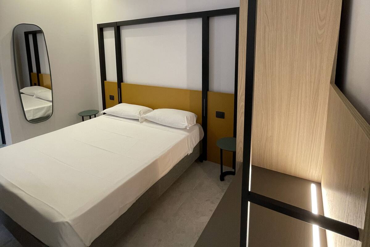 A modern hotel room with a bed, a bedside table, a mirror, and a wooden panel on the wall.