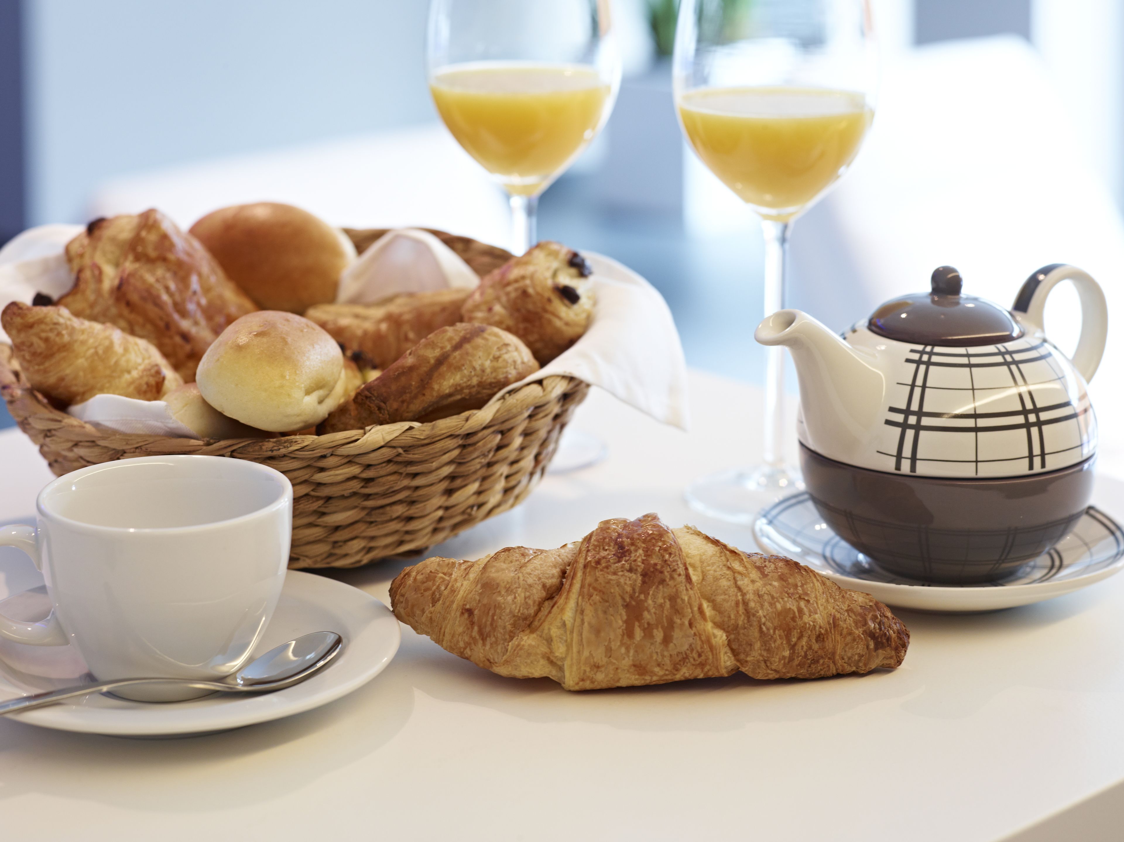 Een ontbijttafel met een mandje croissants, een theepot, een kop en twee glazen jus d’orange.