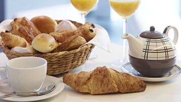 Een ontbijttafel met een mandje croissants, een theepot, een kop en twee glazen jus d’orange.