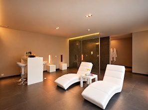 A modern spa room with a glass-enclosed shower, white lounge chairs, a small table with a drink, and a bar area with a bowl of fruit. - Aquabello (Roeselare)