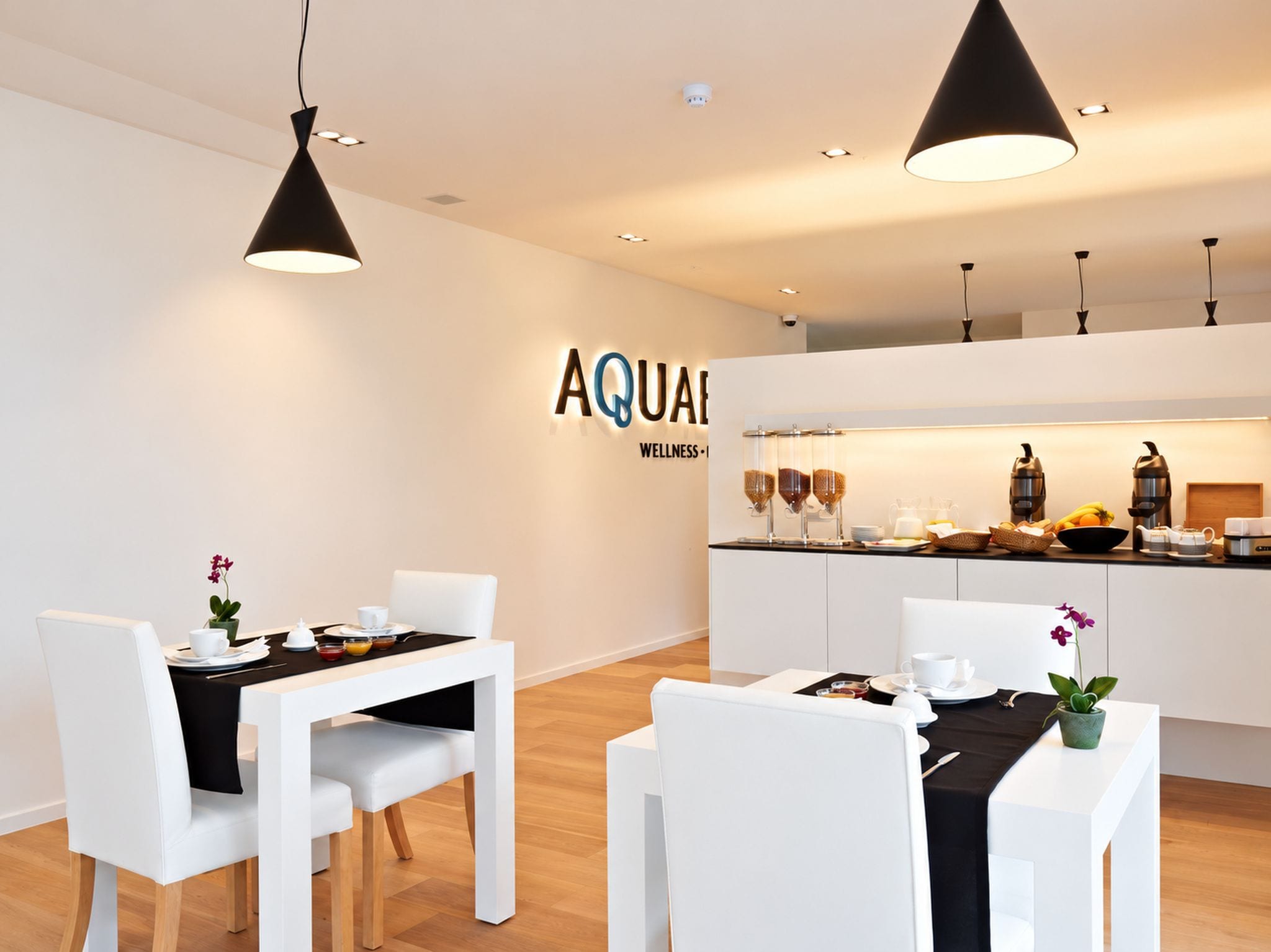 A modern dining area with white chairs and tables, a kitchen in the background, and a sign that reads 'AQUA WELLNESS.'