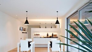 A modern dining area with white furniture, pendant lighting, and a kitchen in the background. - Aquabello (Roeselare)
