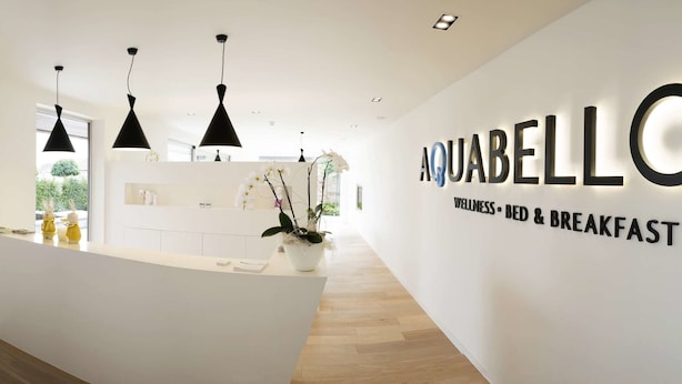 A modern hotel reception area with a white reception desk, black pendant lights, and a sign reading "AQUABELLO WELLNESS BED & BREAKFAST". - Aquabello (Roeselare)