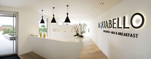 A modern hotel reception area with a white reception desk, black pendant lights, and a sign reading "AQUABELLO WELLNESS BED & BREAKFAST". - Aquabello (Roeselare)