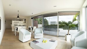 A modern living room with white furniture, a coffee table with magazines, and a large sliding glass door leading to an outdoor area with a view of the sky and trees. - Aquabello (Roeselare)