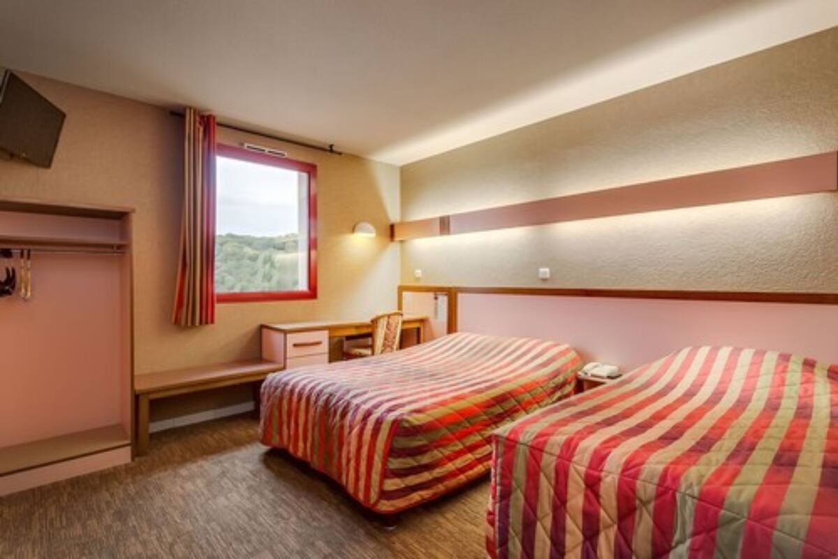 A hotel room with two beds, a desk, a chair, a window with curtains, and a headboard with built-in lighting.