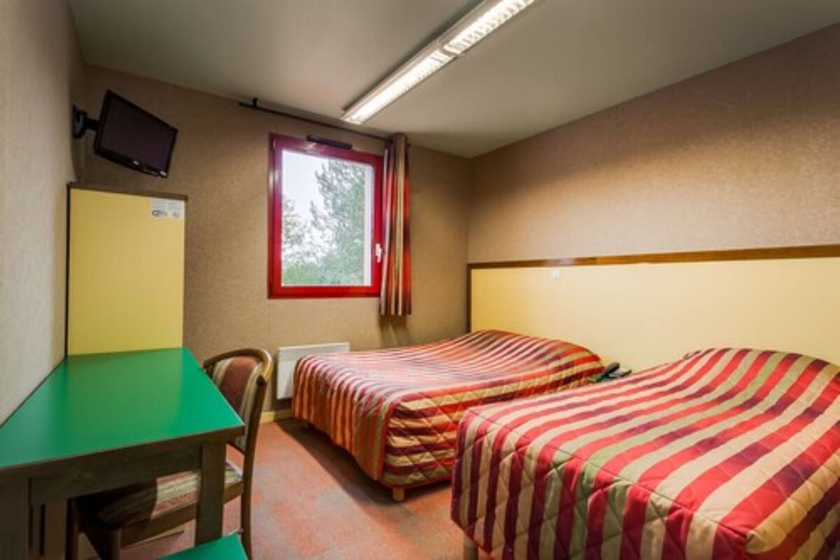 A small room with two single beds, a desk, a chair, a television mounted on the wall, and a window with curtains.