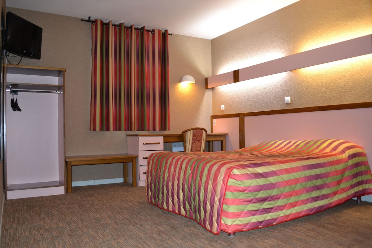 A hotel room with a bed, a desk with a chair, a wardrobe, and a television mounted on the wall.