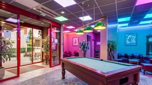 Une table de billard se trouve dans une pièce dotée d’un tapis à motifs, de portes vitrées donnant sur l’extérieur et d’un coin salon avec des chaises et une table.