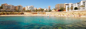 Una playa con agua turquesa cristalina, orilla de arena y edificios al fondo.