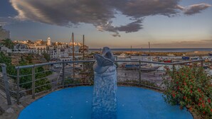 Une ville côtière avec une sculpture imposante au premier plan, un port avec des bateaux et des bâtiments en arrière-plan.
