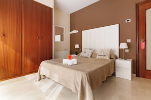 Un dormitorio con un armario grande, una cama con cubrecama color beige, dos mesitas de noche con lámparas y una puerta con cerradura de seguridad.