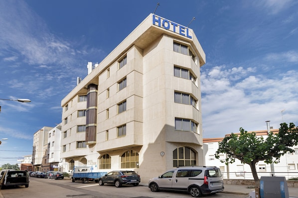 Un hotel con un cartel que indica que es un hotel. Tiene varios pisos con ventanas y un techo plano. Hay coches aparcados frente al hotel.