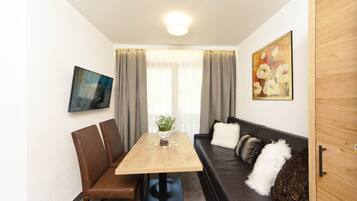 Uma sala de estar moderna com uma mesa de jantar de madeira, cadeiras de couro marrom, um sofá de couro preto com almofadas decorativas, um quadro na parede e uma janela com cortinas leves.