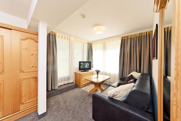A compact living space with a wooden wardrobe, a television on a wooden cabinet, a sofa with a white throw, and a small table with a vase. - Waldpark (Samnaun)