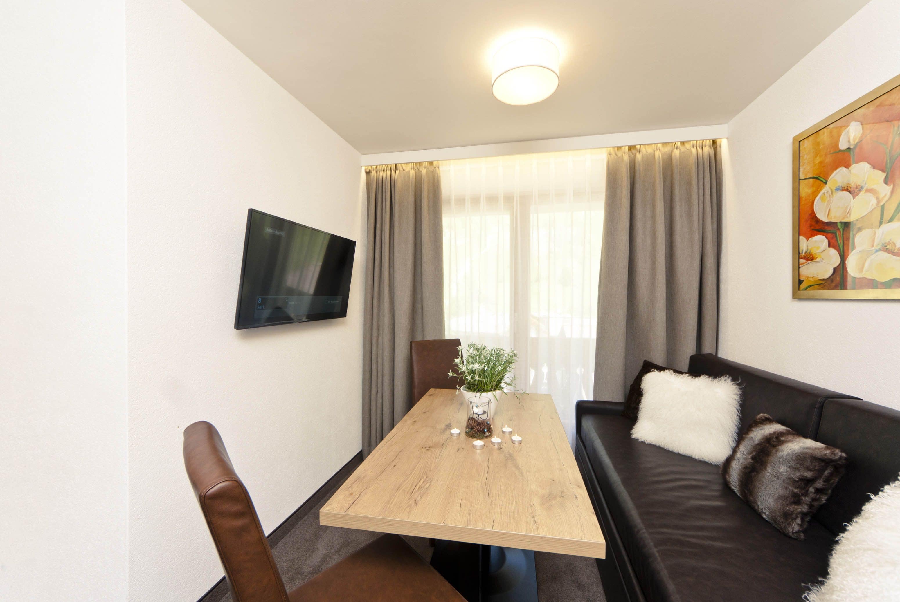 Una sala de estar moderna con una mesa de comedor de madera, un sofá de cuero negro, un televisor montado en la pared y un cuadro colgado.