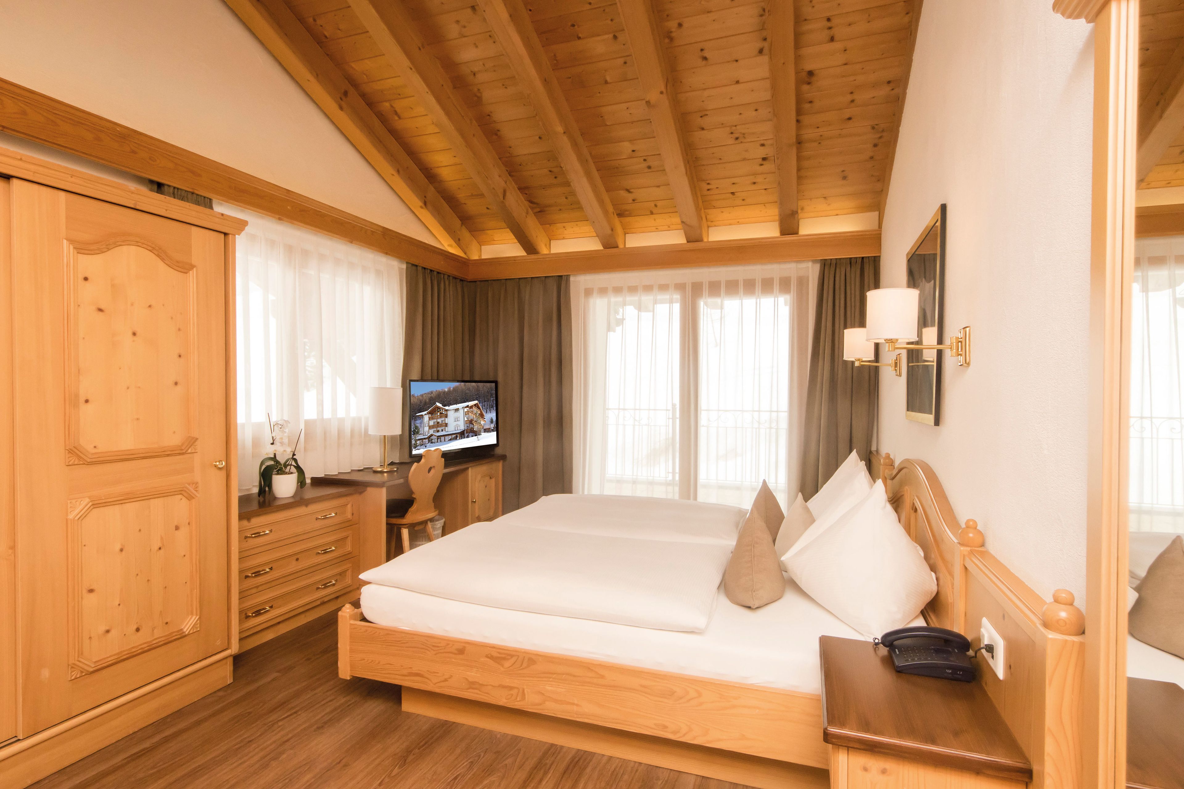 A hotel room with a wooden ceiling, a bed with white linens, a television on a wooden dresser, and a window with curtains.