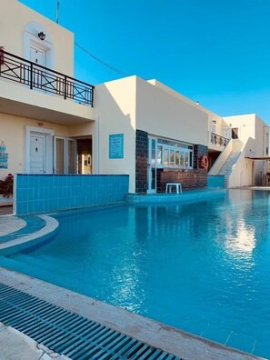 A swimming pool with a clear blue water surface, a white building with a balcony, and a lifebuoy hanging on the wall. - Manine Apartments (Kos)